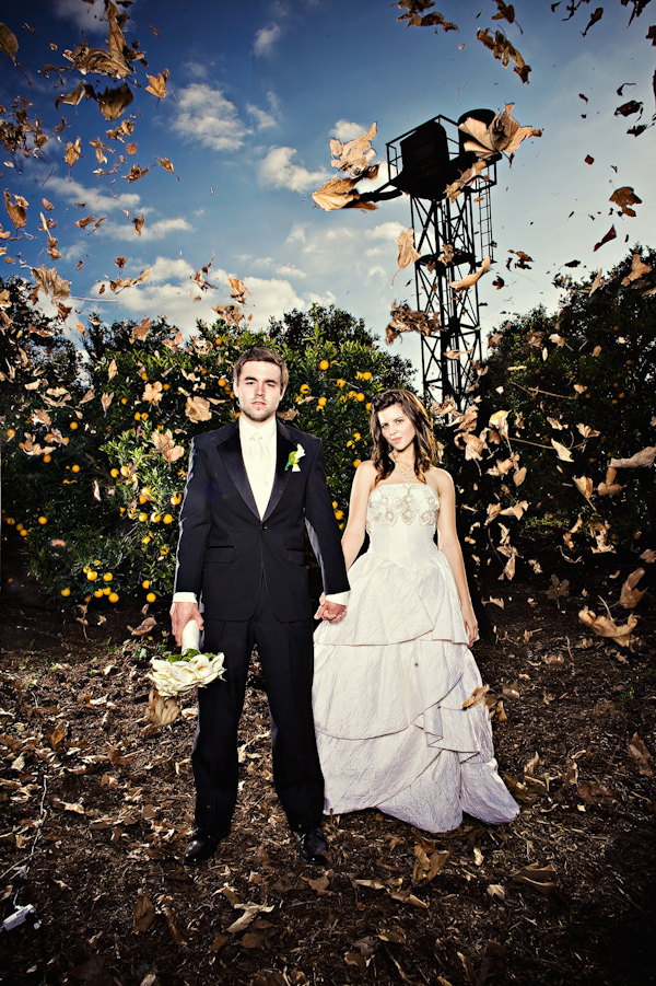 Bride in champagne layered ball gown style wedding dress and groom in black suit with champagne tie and ivory boutonniere holding the bride's ivory bouquet standing in an orchard with autumn leaves blowing all around them - photo by Orange County based wedding photographers Mark Brooke